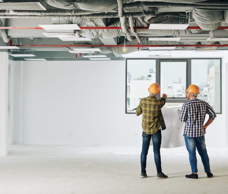 Young construction engineers in plaid shirts and hardhats holding blueprint and looking at city through window