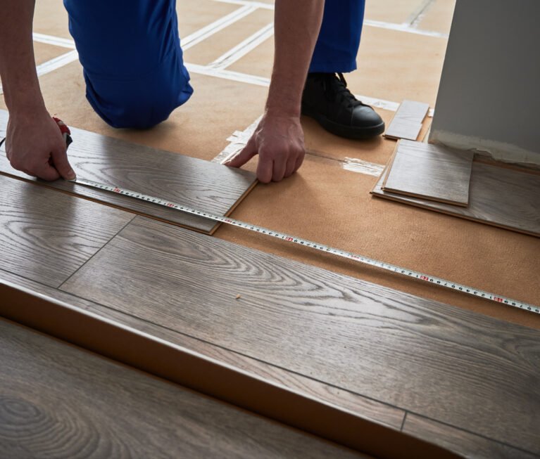 Close up of man construction worker checking distance from wall to laminate board. Male builder using tape measure while installing laminate flooring in apartment under renovation.